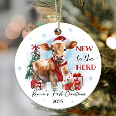 Cow Baby First Christmas Ornament featuring a cute calf in a Santa hat and red scarf, surrounded by gifts and a Christmas tree, with the text New to the Herd and customized with baby’s name and year