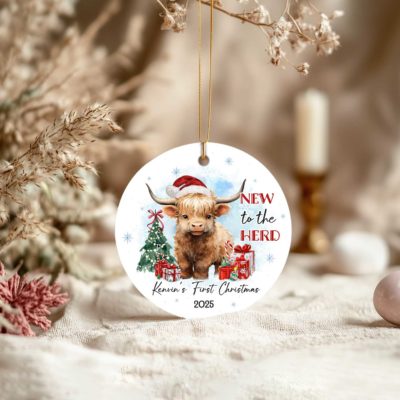 Custom Baby's First Christmas Ornament 2025 featuring a cute Highland cow in a Santa hat and red scarf, with Christmas tree, gifts, and text New to the Herd, customized with baby’s name.
