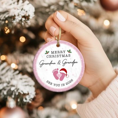 Personalized Baby Announcement Ornament - Christmas Gift for Grandparents featuring baby footprints with a Santa hat and customizable text for names and year, held by hand near a Christmas tree - perfect Christmas pregnancy reveal or gift for grandparents-to-be